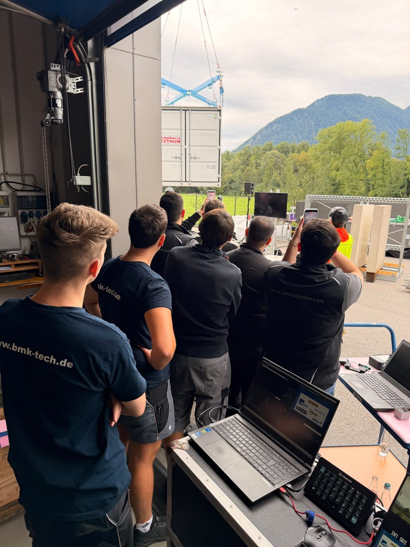 Mitarbeiter und Gäste beobachten am Hallentor einen schwebenden Container am Kran, im Vordergrund Laptop und Stream Deck, im Hintergrund die Berge